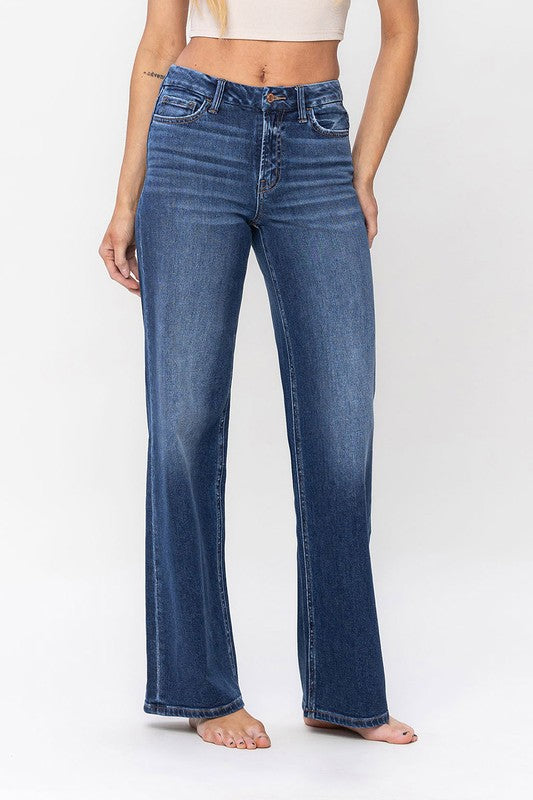 A person stands barefoot against a plain background, wearing a sleeveless top and the High Rise Loose Fit Jeans. These blue wide-leg jeans provide a loose fit that exudes comfort and effortless style.