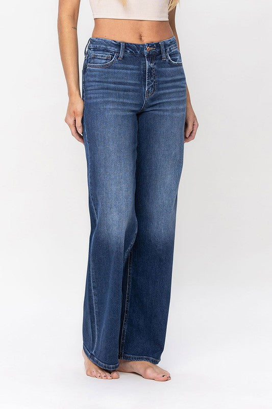 A person stands barefoot against a plain background, wearing a sleeveless top and the High Rise Loose Fit Jeans. These blue wide-leg jeans provide a loose fit that exudes comfort and effortless style.