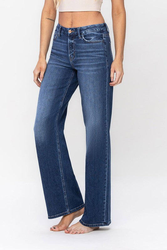 A person stands barefoot against a plain background, wearing a sleeveless top and the High Rise Loose Fit Jeans. These blue wide-leg jeans provide a loose fit that exudes comfort and effortless style.