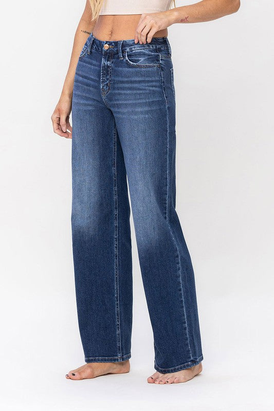 A person stands barefoot against a plain background, wearing a sleeveless top and the High Rise Loose Fit Jeans. These blue wide-leg jeans provide a loose fit that exudes comfort and effortless style.