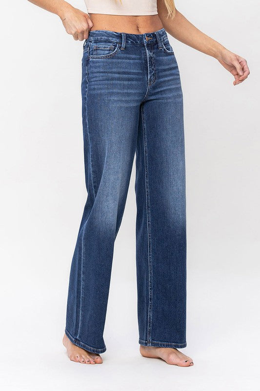 A person stands barefoot against a plain background, wearing a sleeveless top and the High Rise Loose Fit Jeans. These blue wide-leg jeans provide a loose fit that exudes comfort and effortless style.