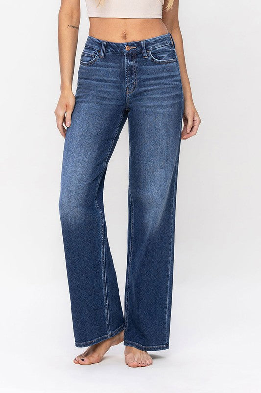 A person stands barefoot against a plain background, wearing a sleeveless top and the High Rise Loose Fit Jeans. These blue wide-leg jeans provide a loose fit that exudes comfort and effortless style.