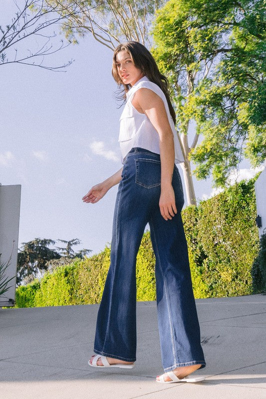 A person stands barefoot against a plain background, wearing a sleeveless top and the High Rise Loose Fit Jeans. These blue wide-leg jeans provide a loose fit that exudes comfort and effortless style.