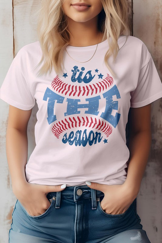 A person wearing the "Tis The Season, Baseball Graphic Tee" and blue jeans, standing against a wooden background.