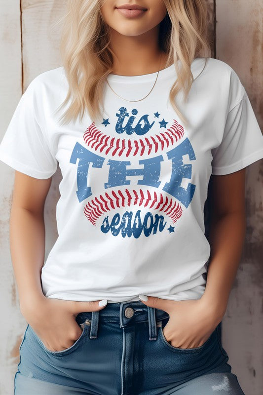 A person wearing the "Tis The Season, Baseball Graphic Tee" and blue jeans, standing against a wooden background.