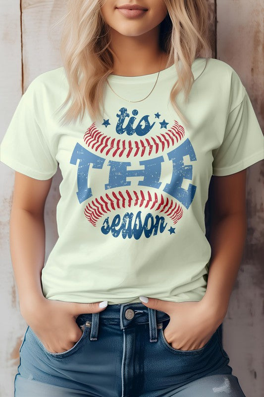 A person wearing the "Tis The Season, Baseball Graphic Tee" and blue jeans, standing against a wooden background.