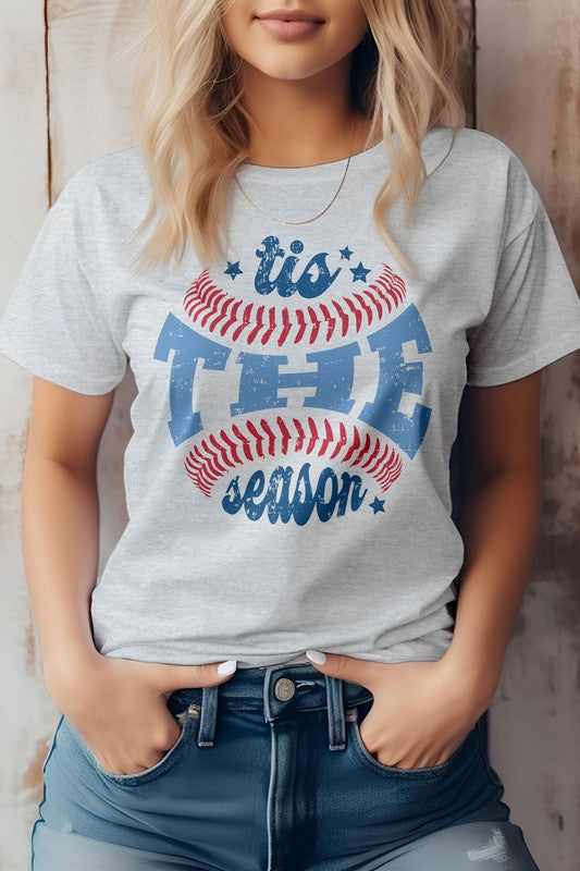 A person wearing the "Tis The Season, Baseball Graphic Tee" and blue jeans, standing against a wooden background.