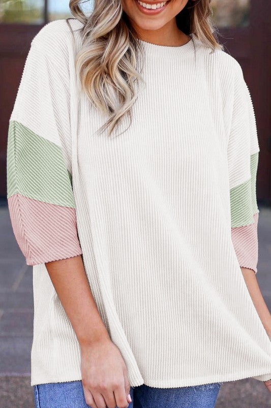 A woman with long blonde hair is seen wearing the Ribbed Stripe Drop Shoulder Half Sleeve Top Blouse, which features a relaxed fit and a color block design with green and pink sleeve accents. She pairs it with blue jeans and stands against a light pink wall, accessorized with gold necklaces and a bracelet.