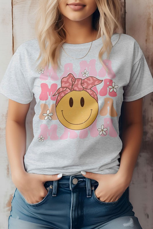 A woman is wearing the MAMA, Smiley, Graphic Tee in light pink, featuring a smiley face and the word "mama" repeatedly written around it. She poses with her hands in her jean pockets, making it a perfect outfit for Mother's Day. The unisex fit adds a stylish touch to any wardrobe.