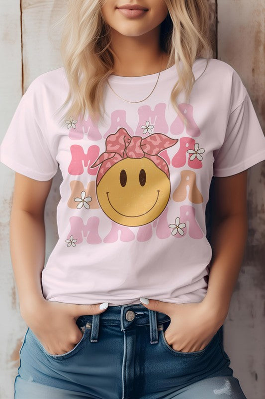 A woman is wearing the MAMA, Smiley, Graphic Tee in light pink, featuring a smiley face and the word "mama" repeatedly written around it. She poses with her hands in her jean pockets, making it a perfect outfit for Mother's Day. The unisex fit adds a stylish touch to any wardrobe.