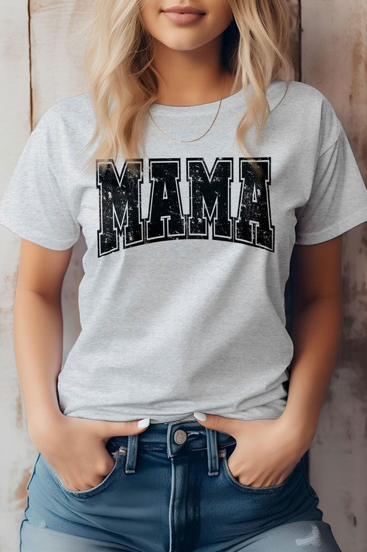 A person wearing the Mama Retro Varsity Graphic Tee in white, showcasing the word "MAMA" in large, distressed pink letters, stands against a wooden background with their hands in the front pockets of blue jeans. This look is perfect for Mother's Day.