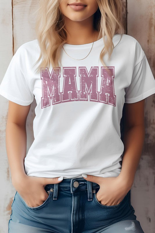 A person wearing the Mama Retro Varsity Graphic Tee in white, showcasing the word "MAMA" in large, distressed pink letters, stands against a wooden background with their hands in the front pockets of blue jeans. This look is perfect for Mother's Day.