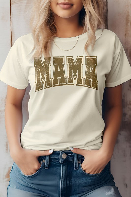 A person wearing the Mama Retro Varsity Graphic Tee in white, showcasing the word "MAMA" in large, distressed pink letters, stands against a wooden background with their hands in the front pockets of blue jeans. This look is perfect for Mother's Day.