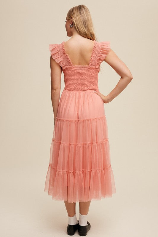 A woman in a Smocked Ruffle Tiered Mesh Midi Maxi Dress, featuring mauve tulle layers with smocked ruffles and ruffled straps, poses against a plain background. She is accessorized with long earrings and silver heels.