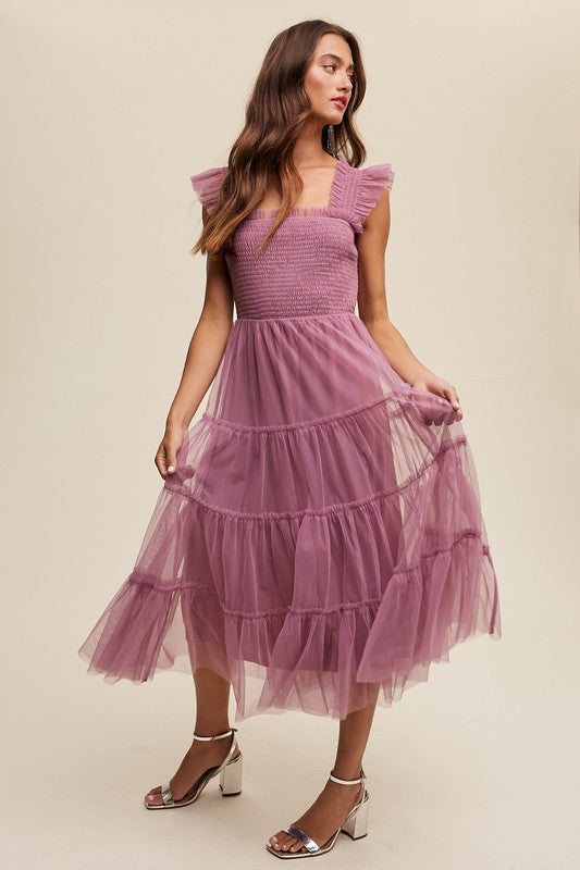 A woman in a Smocked Ruffle Tiered Mesh Midi Maxi Dress, featuring mauve tulle layers with smocked ruffles and ruffled straps, poses against a plain background. She is accessorized with long earrings and silver heels.