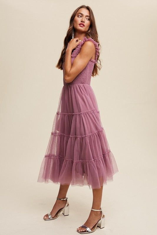 A woman in a Smocked Ruffle Tiered Mesh Midi Maxi Dress, featuring mauve tulle layers with smocked ruffles and ruffled straps, poses against a plain background. She is accessorized with long earrings and silver heels.