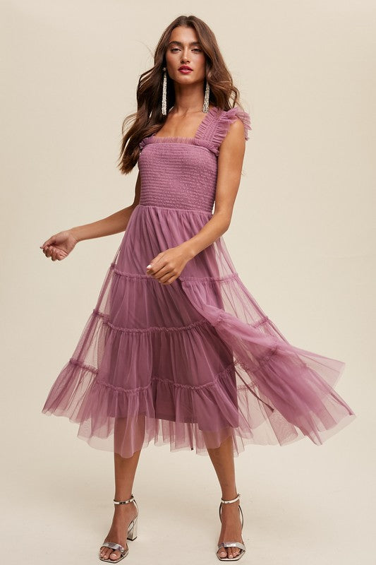 A woman in a Smocked Ruffle Tiered Mesh Midi Maxi Dress, featuring mauve tulle layers with smocked ruffles and ruffled straps, poses against a plain background. She is accessorized with long earrings and silver heels.