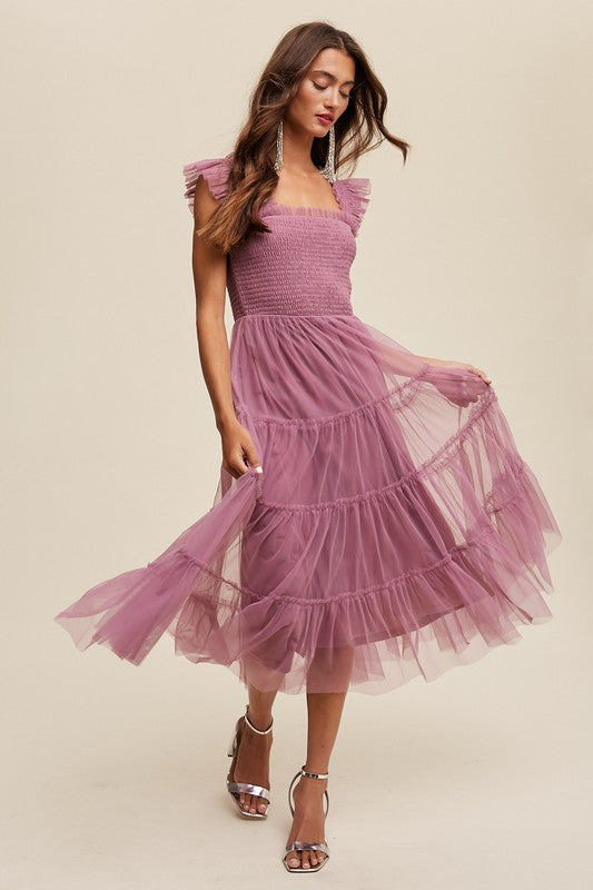 A woman in a Smocked Ruffle Tiered Mesh Midi Maxi Dress, featuring mauve tulle layers with smocked ruffles and ruffled straps, poses against a plain background. She is accessorized with long earrings and silver heels.