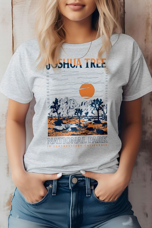 A person wearing an eco-friendly beige graphic tee featuring a design of Joshua Tree National Park with desert scenery, mountains, and an orange sun. The text on the shirt reads "Joshua Tree in Desert Mountain.