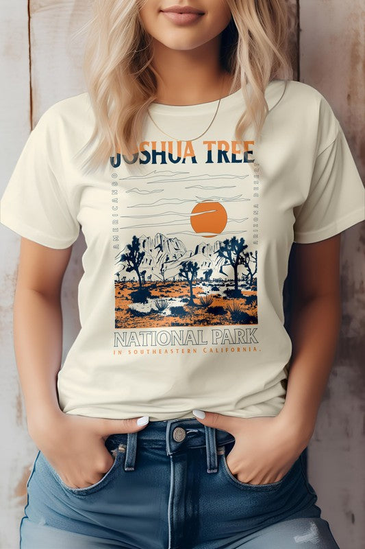 A person wearing an eco-friendly beige graphic tee featuring a design of Joshua Tree National Park with desert scenery, mountains, and an orange sun. The text on the shirt reads "Joshua Tree in Desert Mountain.