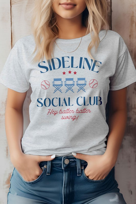 A person is wearing the Sideline Social Club Baseball Graphic Tee, displaying the words "Hey batter batter swing!" alongside images of two baseballs and three director's chairs. This white T-shirt features a unisex fit, making it an ideal choice for any baseball fan.