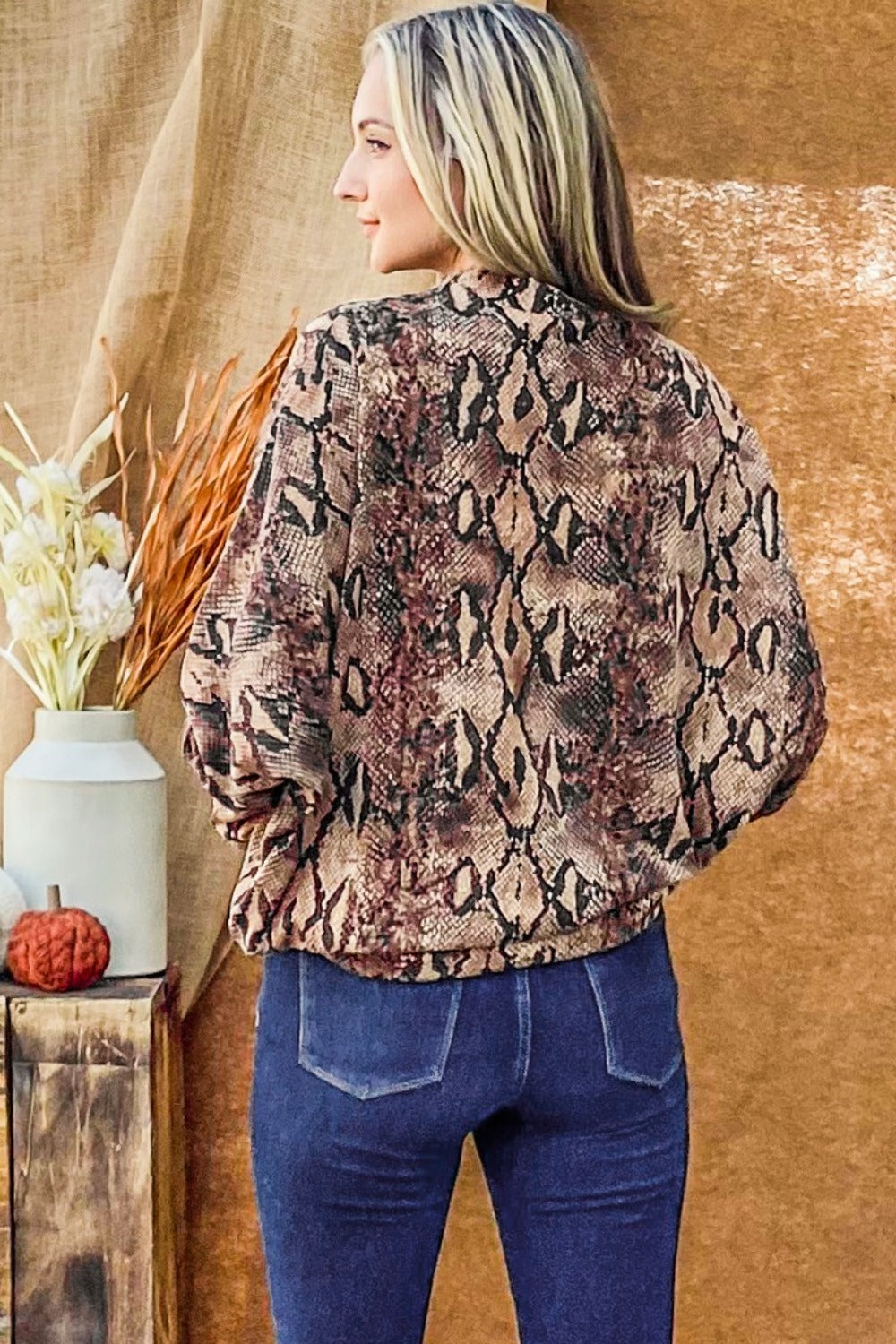 A woman wearing the And The Why Choker Neck Dolman Sleeve Snake Print Top paired with high-waisted jeans, smiling and posing in front of a brown background with decorative elements.