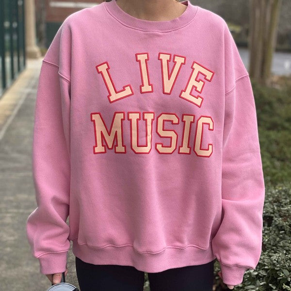 The Live Music Lover Oversized Sweatshirt, in a cozy and stylish pink design with "LIVE MUSIC LOVER" printed in yellow and white letters, is displayed on a wooden hanger. An arm is visible holding the hanger, emphasizing the sweatshirt's oversized fit.