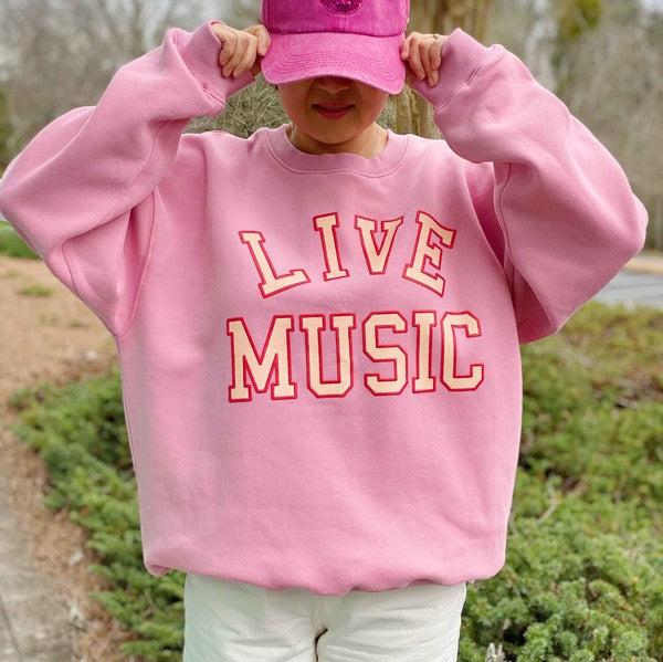 The Live Music Lover Oversized Sweatshirt, in a cozy and stylish pink design with "LIVE MUSIC LOVER" printed in yellow and white letters, is displayed on a wooden hanger. An arm is visible holding the hanger, emphasizing the sweatshirt's oversized fit.