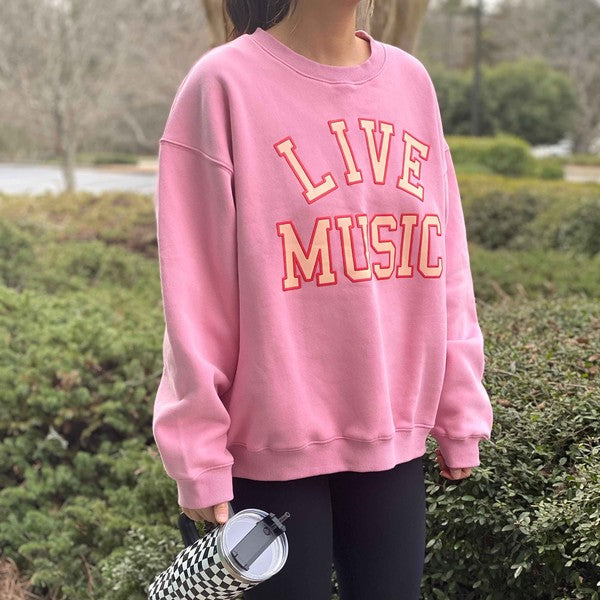 The Live Music Lover Oversized Sweatshirt, in a cozy and stylish pink design with "LIVE MUSIC LOVER" printed in yellow and white letters, is displayed on a wooden hanger. An arm is visible holding the hanger, emphasizing the sweatshirt's oversized fit.