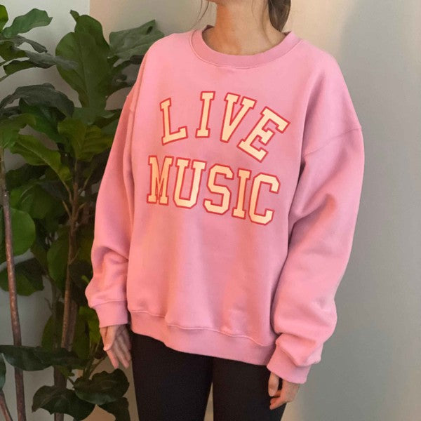 The Live Music Lover Oversized Sweatshirt, in a cozy and stylish pink design with "LIVE MUSIC LOVER" printed in yellow and white letters, is displayed on a wooden hanger. An arm is visible holding the hanger, emphasizing the sweatshirt's oversized fit.