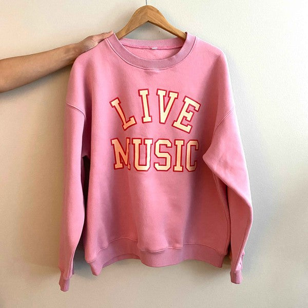 The Live Music Lover Oversized Sweatshirt, in a cozy and stylish pink design with "LIVE MUSIC LOVER" printed in yellow and white letters, is displayed on a wooden hanger. An arm is visible holding the hanger, emphasizing the sweatshirt's oversized fit.