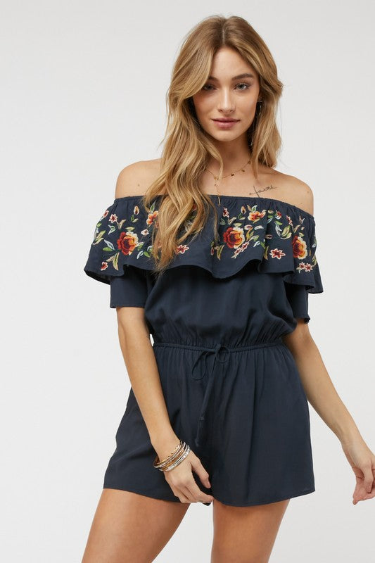 Wearing the stylish Floral Embroidered Off Shoulder Romper, a woman poses gracefully, showcasing its ruffle detail and making it an ideal addition to any summer wardrobe.