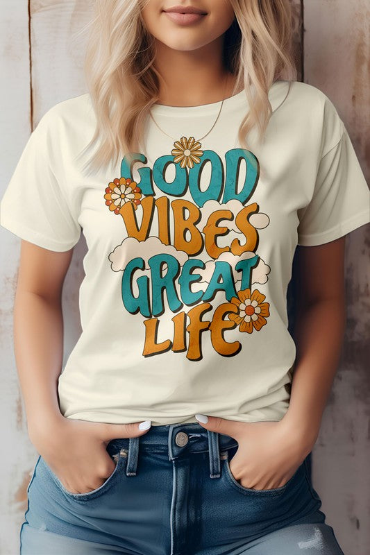 A person wearing the Good Vibes Great Life, Retro Vintage Graphic Tee, featuring colorful text and floral designs on a white background, stands with hands in jeans pockets, showcasing an eco-friendly material that aligns perfectly with the hippie style.