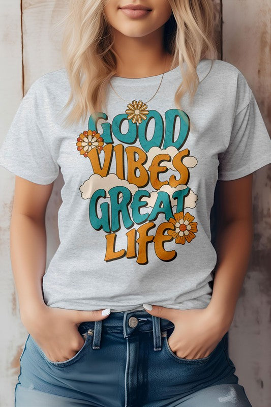 A person wearing the Good Vibes Great Life, Retro Vintage Graphic Tee, featuring colorful text and floral designs on a white background, stands with hands in jeans pockets, showcasing an eco-friendly material that aligns perfectly with the hippie style.