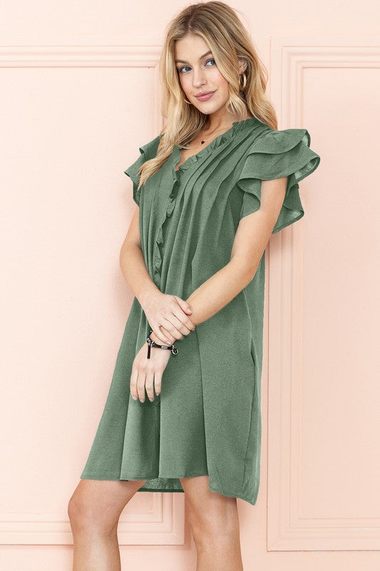 Woman in a green Ruffle Sleeve V Neck dress with pockets, standing against a peach-colored wall, smiling with hands on hips.