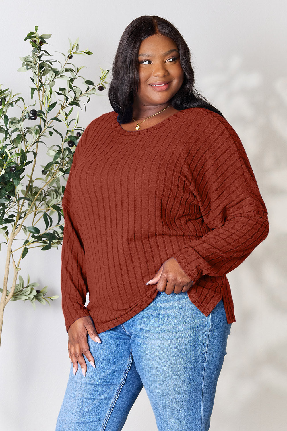 A person wearing the Basic Bae Full Size Ribbed Round Neck Slit Knit Top in blue and beige pants stands and smiles in front of a leafy plant.