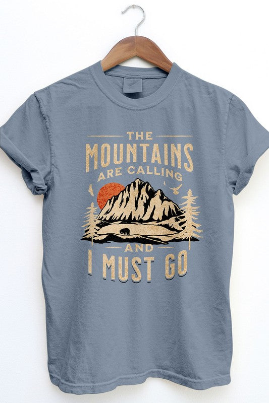 The Mountains are Calling Garment Dye Tee, in a vintage wash dark gray, hangs on a wooden hanger displaying an outdoor design with mountains, trees, and a bear. The shirt features the text "The mountains are calling and I must go" and offers unisex appeal while capturing the essence of adventure in timeless style.