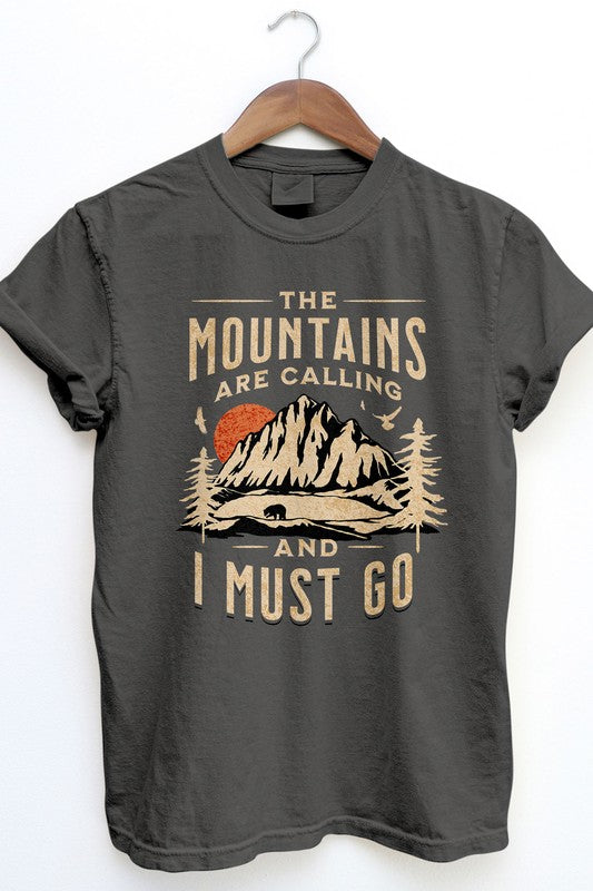 The Mountains are Calling Garment Dye Tee, in a vintage wash dark gray, hangs on a wooden hanger displaying an outdoor design with mountains, trees, and a bear. The shirt features the text "The mountains are calling and I must go" and offers unisex appeal while capturing the essence of adventure in timeless style.