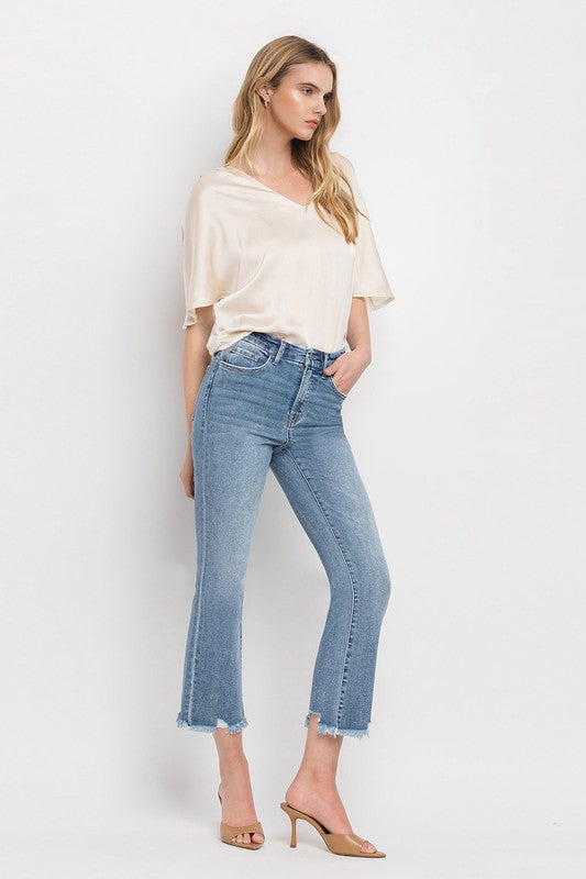 A woman stands against a plain white background wearing a beige blouse, High Rise Crop Flare Jeans made from comfort stretch denim with a raw hem, and tan high heels. She has long, wavy hair and is looking at the camera with a neutral expression.