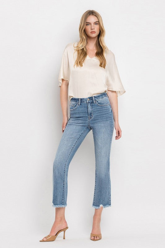 A woman stands against a plain white background wearing a beige blouse, High Rise Crop Flare Jeans made from comfort stretch denim with a raw hem, and tan high heels. She has long, wavy hair and is looking at the camera with a neutral expression.