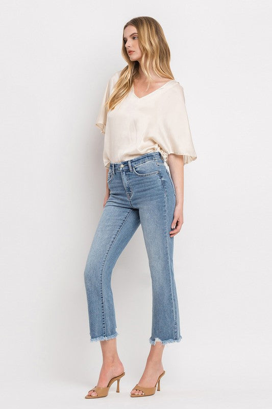 A woman stands against a plain white background wearing a beige blouse, High Rise Crop Flare Jeans made from comfort stretch denim with a raw hem, and tan high heels. She has long, wavy hair and is looking at the camera with a neutral expression.