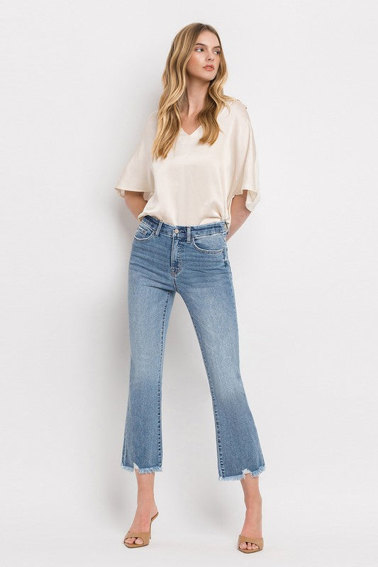 A woman stands against a plain white background wearing a beige blouse, High Rise Crop Flare Jeans made from comfort stretch denim with a raw hem, and tan high heels. She has long, wavy hair and is looking at the camera with a neutral expression.