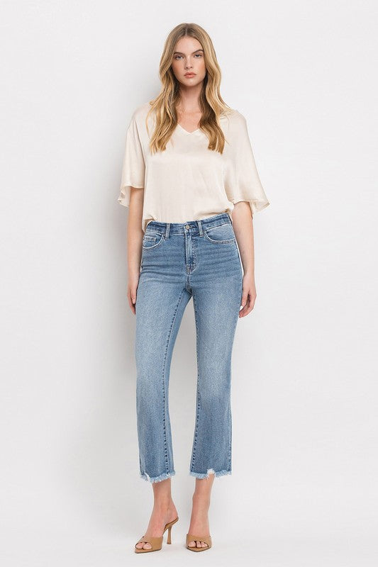 A woman stands against a plain white background wearing a beige blouse, High Rise Crop Flare Jeans made from comfort stretch denim with a raw hem, and tan high heels. She has long, wavy hair and is looking at the camera with a neutral expression.