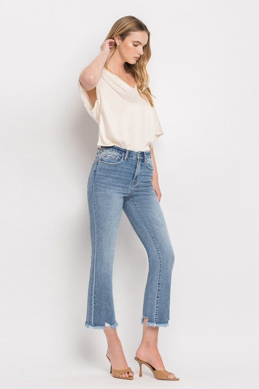 A woman stands against a plain white background wearing a beige blouse, High Rise Crop Flare Jeans made from comfort stretch denim with a raw hem, and tan high heels. She has long, wavy hair and is looking at the camera with a neutral expression.