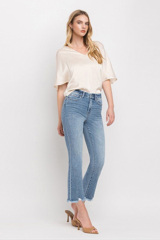 A woman stands against a plain white background wearing a beige blouse, High Rise Crop Flare Jeans made from comfort stretch denim with a raw hem, and tan high heels. She has long, wavy hair and is looking at the camera with a neutral expression.