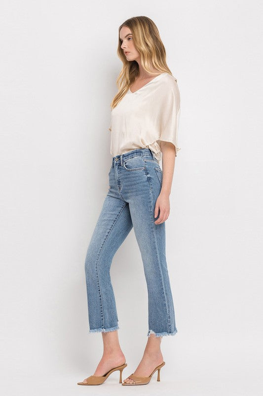 A woman stands against a plain white background wearing a beige blouse, High Rise Crop Flare Jeans made from comfort stretch denim with a raw hem, and tan high heels. She has long, wavy hair and is looking at the camera with a neutral expression.