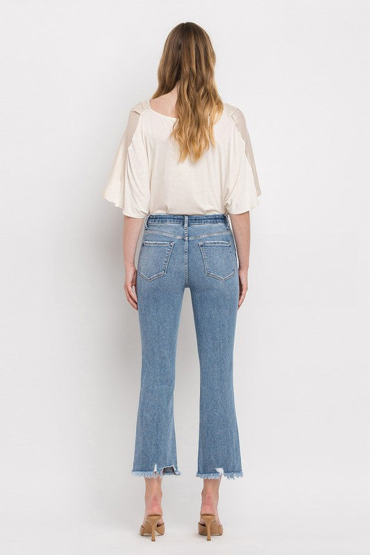 A woman stands against a plain white background wearing a beige blouse, High Rise Crop Flare Jeans made from comfort stretch denim with a raw hem, and tan high heels. She has long, wavy hair and is looking at the camera with a neutral expression.