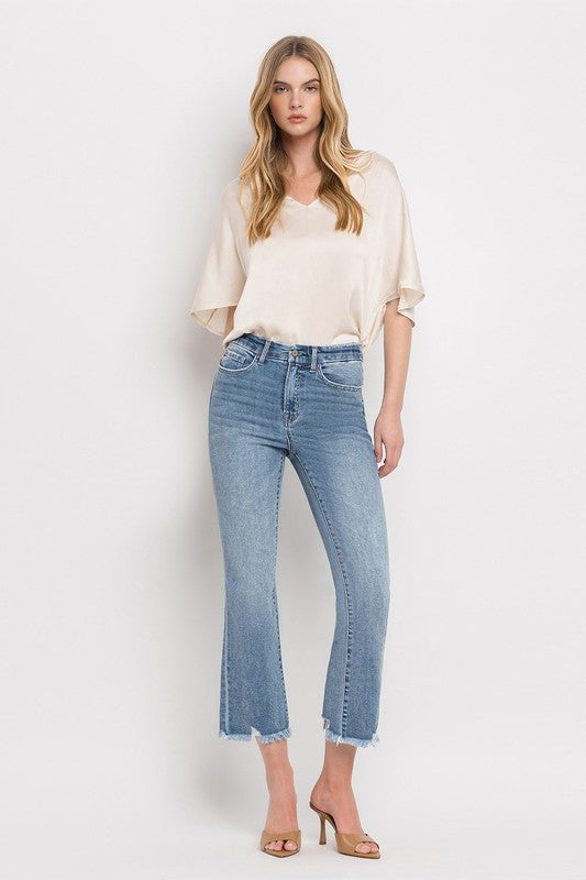 A woman stands against a plain white background wearing a beige blouse, High Rise Crop Flare Jeans made from comfort stretch denim with a raw hem, and tan high heels. She has long, wavy hair and is looking at the camera with a neutral expression.