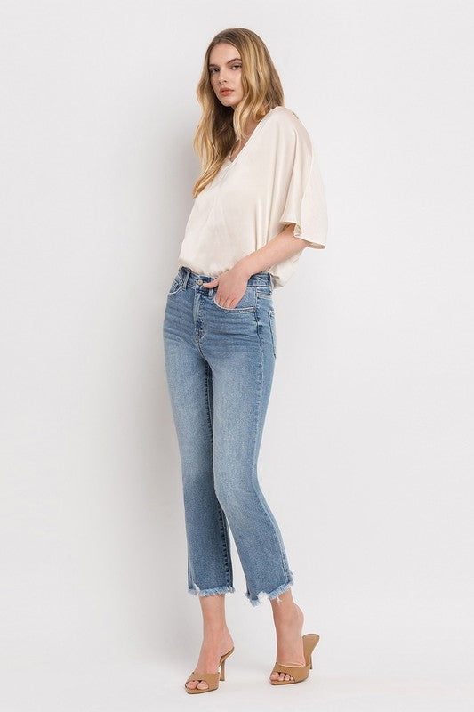 A woman stands against a plain white background wearing a beige blouse, High Rise Crop Flare Jeans made from comfort stretch denim with a raw hem, and tan high heels. She has long, wavy hair and is looking at the camera with a neutral expression.