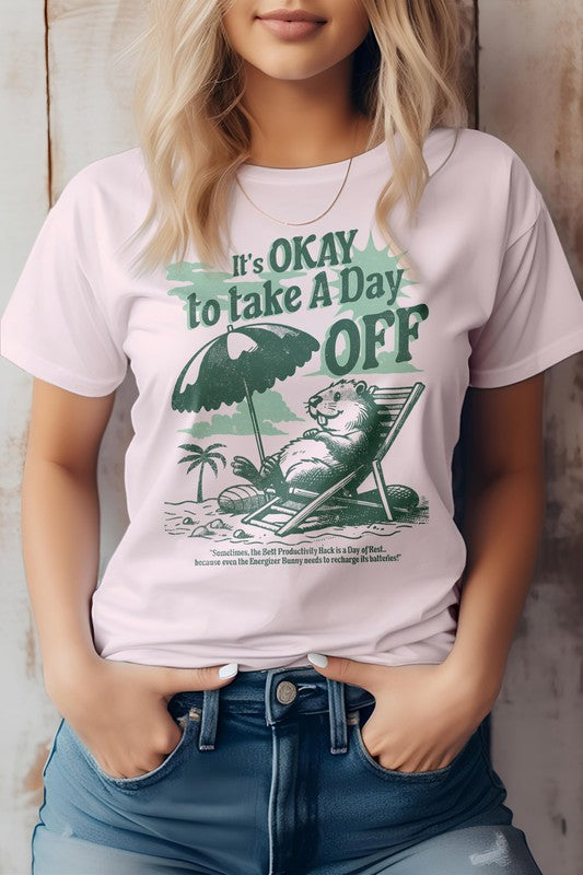 A person wearing the Day Off, Retro Vintage Graphic Tee in light green, featuring an illustration of a beaver relaxing on a beach under an umbrella. Made from eco-friendly materials, this shirt proudly displays the message "It's OKAY to Take A Day OFF" in bold letters.